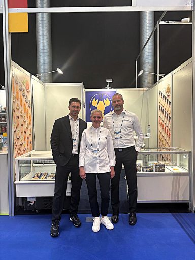 Drei Personen in formeller Kleidung stehen vor dem Christian Goedeken Messestand mit bunten Produkten auf der PLMA.