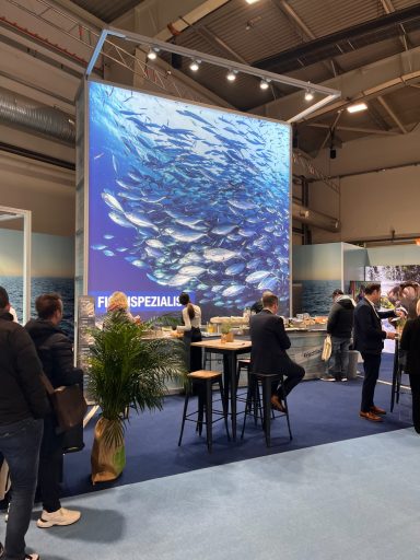 Messestand mit großem Ozean-Motiv und Besuchern, die sich um Stehtische versammeln.